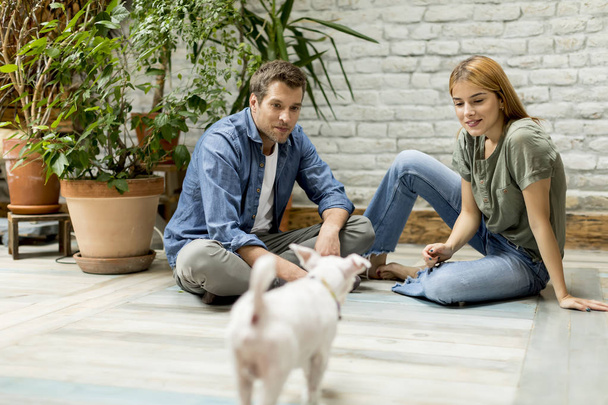 schönes junges Paar sitzt am rustikalen Wohnzimmerboden und spielt mit niedlichem weißen Hund - Foto, Bild