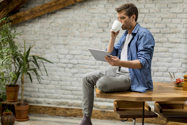 schöner junger Mann mit digitalem Tablet und Kaffee trinken, während er auf dem Tisch im rustikalen Zimmer sitzt - Foto, Bild