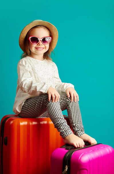 Vertical shot of smiling little girl in straw hat and sunglasses sitting on suitcases, portrait of beautiful child going on a journey, studio shot over isolated blue isolated. - Foto, Imagen