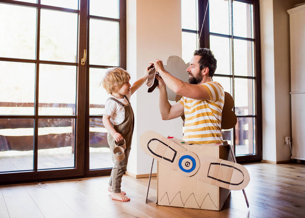 A toddler boy and father with carton plane playing indoors at home, flying concept. - Zdjęcie, obraz