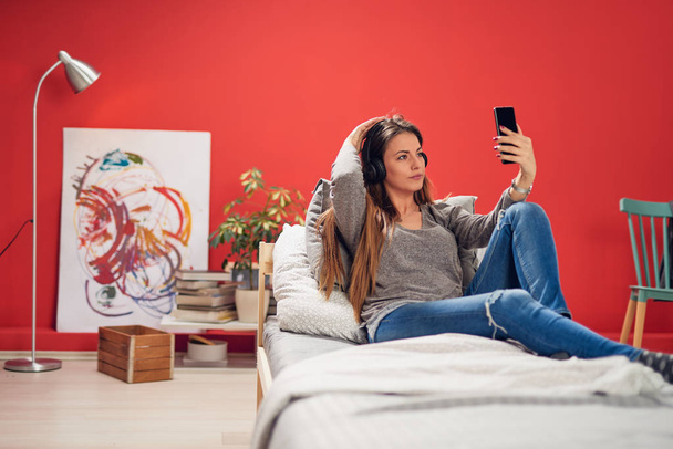 Beautiful smiling Caucasian brunette dressed casual and with haedphones on ears sitting on bed in bedroom and taking selfie. - Фото, изображение