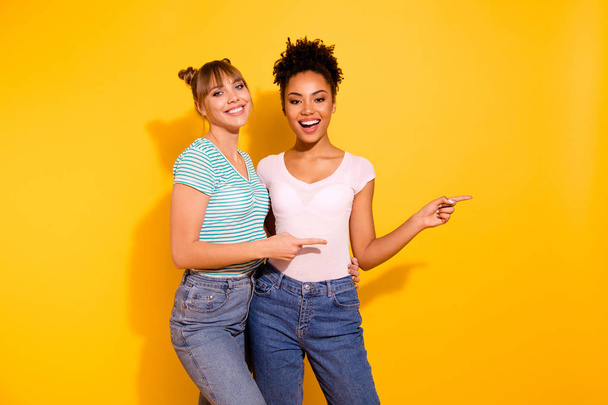 Close up photo funny beautiful two people she her model lady diversity raise index fingers side empty space amazed toothy wear casual white striped t-shirt clothes isolated yellow bright background - 写真・画像