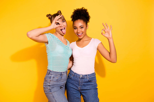 Close up photo funky beautiful she her lady hug embrace cuddle different buddies hold hands arms okey symbol tongue out mouth wear casual white striped t-shirt outfit isolated yellow bright background - Φωτογραφία, εικόνα