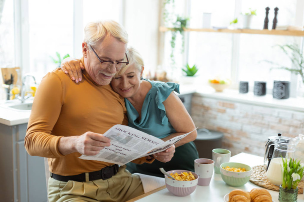 Smiling nice-appealing aged white-haired married couple reading a newspaper together - Foto, afbeelding
