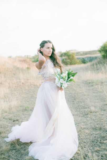 Beautiful young bride with long hair in a wedding dress with a bouquet in the summer in the field - Foto, afbeelding