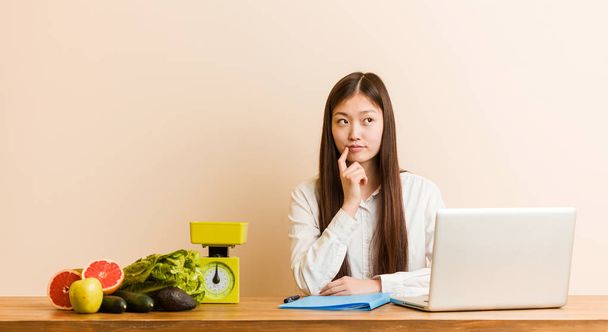 Jovem nutricionista mulher chinesa trabalhando com seu laptop olhando para o lado com expressão duvidosa e cética
. - Foto, Imagem