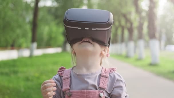 Little caucasian girl 4-5 years using VR glasses headset on nature. Children in pink sundress wearing virtual reality glasses with fun and surprising face standing outdoor. Close up shot - Felvétel, videó