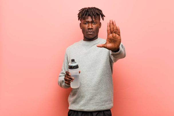 Joven fitness hombre negro sosteniendo una botella de agua de pie con la mano extendida mostrando señal de stop, lo que le impide
. - Foto, imagen