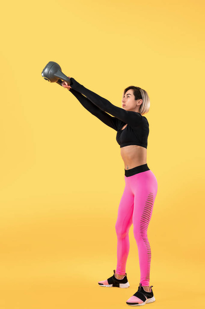Fitness woman in fashionable pink and black sportswear work out with kettlebell on yellow background. - Zdjęcie, obraz