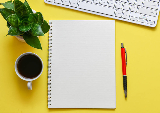 Ansicht des Arbeitsplatzes mit leerem Notizblock, Stift, Tastatur und grüner Pflanze auf gelbem Hintergrund  - Foto, Bild