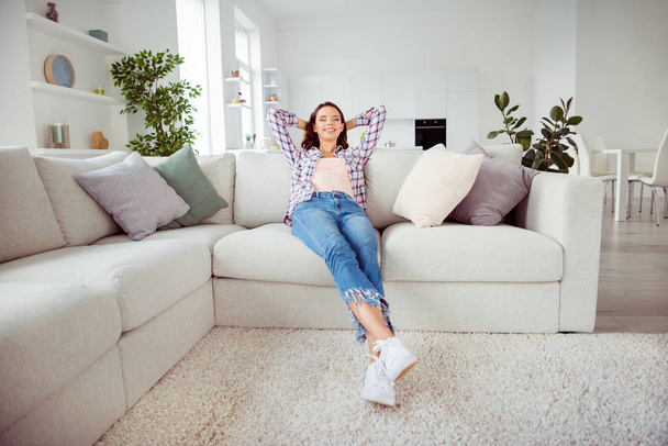 Full length body size photo amazing beautiful she her lady eyes closed overjoyed hands arms behind head wear checkered plaid shirt jeans denim sit comfort divan house loft living room indoors - 写真・画像