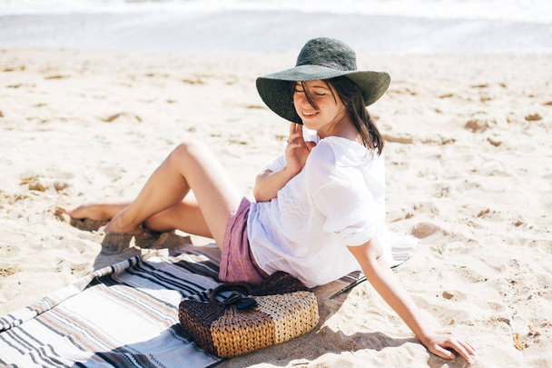Stylish hipster girl in hat sitting on beach with straw bag and  - Fotografie, Obrázek