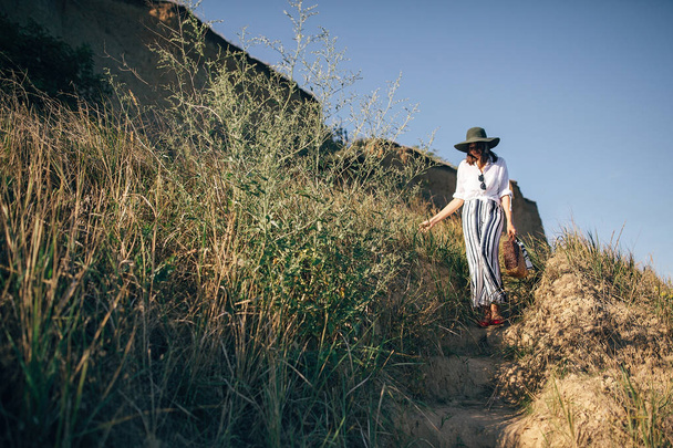 Stylowa dziewczyna Boho w kapeluszu chodzenie na piaszczystej klifie z trawą w pobliżu  - Zdjęcie, obraz