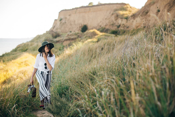 Stylish boho girl in hat walking at sandy cliff with grass near  - Фото, зображення