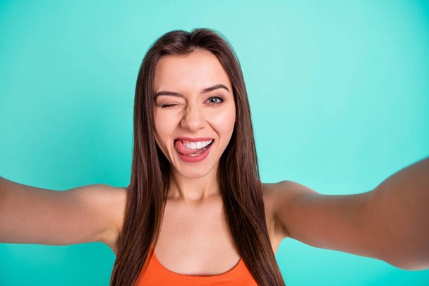 Close up photo beautiful amazing her she lady make take selfies tell speak say skype tongue out mouth blink one eye flirty wear casual orange tank-top isolated bright blue teal turquoise background - 写真・画像