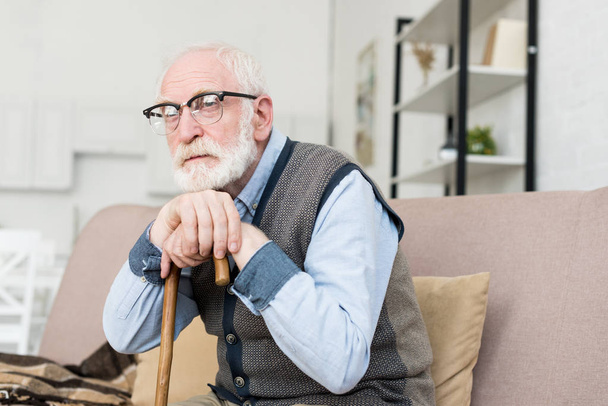 Bearded and grey haired man sitting in bright room, putting hands on walking stick - Photo, Image