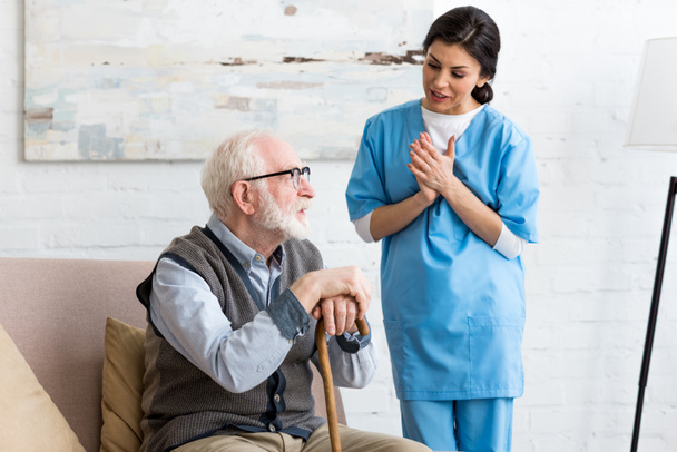 Nurse talking to bearded senior man, standing inside bright room - Фото, изображение