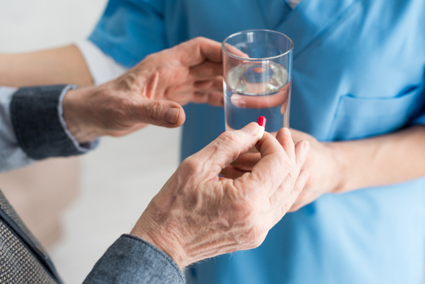 Обрезанный вид пожилого человека с таблетками в руке, в то время как доктор дает стакан с водой для пациента
 - Фото, изображение