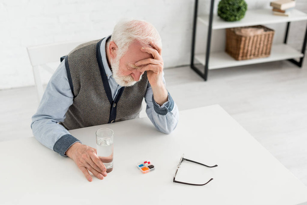 Vue d'angle élevé de l'homme âgé bouleversé assis derrière la table avec des pilules, tenant la main sur la tête
 - Photo, image