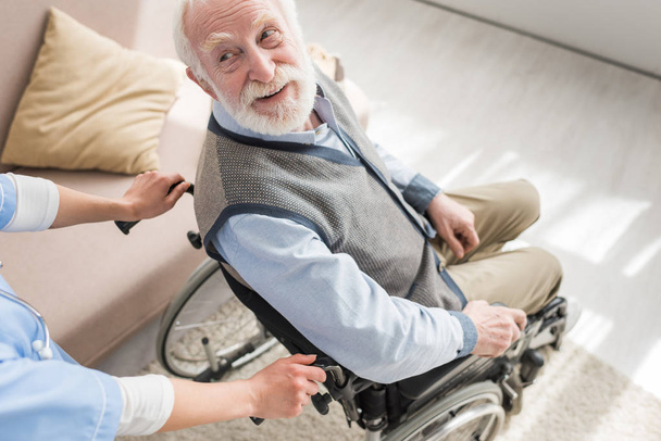 High angle view of disabled and cheerful gray haired man in wheelchair looking at nurse - 写真・画像