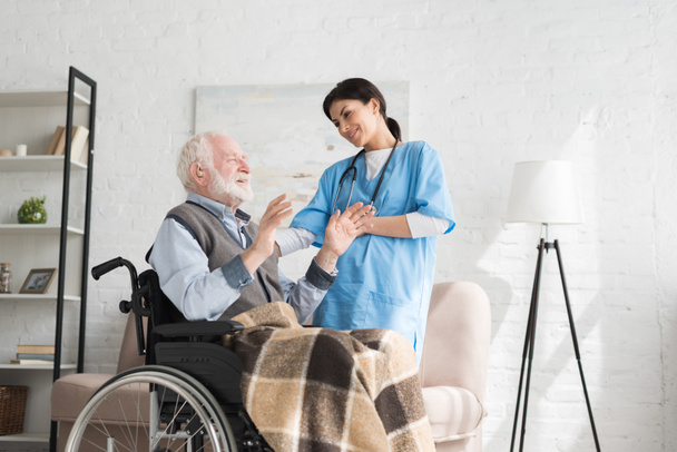 Nurse talking to disabled and happy grey haired man in wheelchair - Foto, imagen