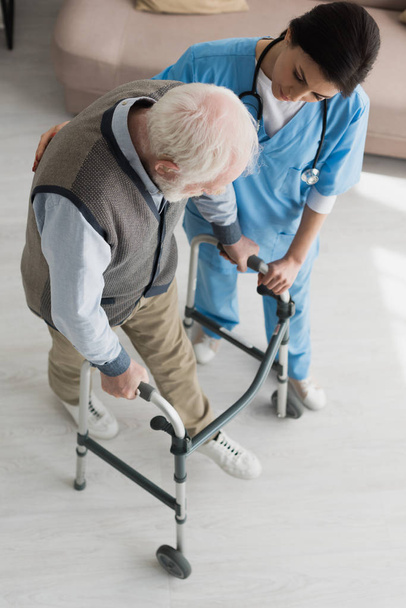 Высокий угол обзора врача, помогающего старику ходить, выздоравливая после травмы
 - Фото, изображение