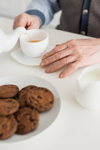 Vue recadrée de l'homme âgé main sur table blanche avec tasse de thé et biscuits
 - Photo, image
