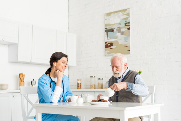 Happy doctor siting on kitchen behind table with senior man - Фото, изображение