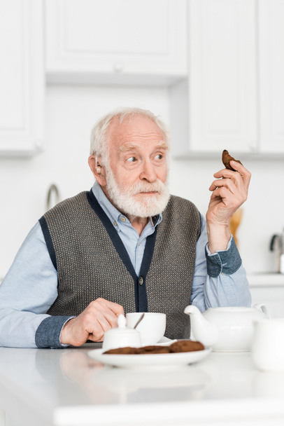 Curious grey haired man sitting on kitchen with cookie in hand, and looking away - Φωτογραφία, εικόνα