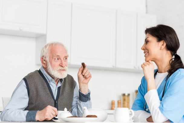 Cheerful grey haired man sitting on kitchen with cookie in hand, and looking at nurse - Foto, imagen