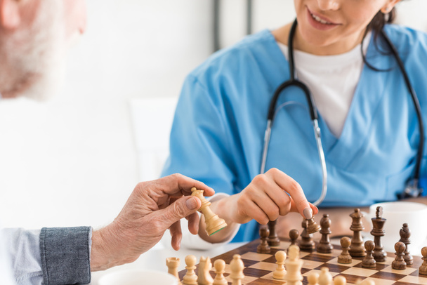 Vista ritagliata di infermiera e uomo anziano seduto in cucina, e giocare a scacchi
 - Foto, immagini