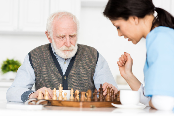 Selective focus on retired man sitting on kitchen, and playing in chess with nurse - Photo, Image