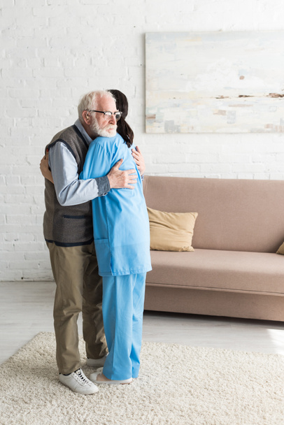 Calm senior man hugging with nurse, standing in bright room - 写真・画像