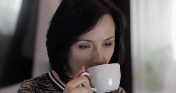 カップからコーヒーを飲んで微笑んで飲むかわいい若いブルネットの女性の肖像画 - 映像、動画