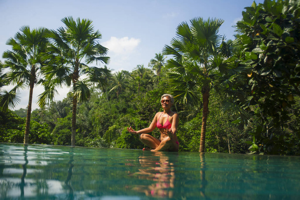 attractive and happy middle aged Asian Indonesian woman in bikini relaxed at infinity pool of exotic resort in tropical jungle sitting in lotus yoga position meditating  - Foto, Bild