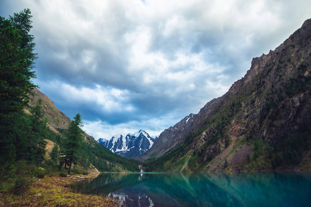 Wonderful mountain lake with view on giant glacier. Amazing huge mountains with conifer forest. Larch tree on water edge. Morning landscape of majestic nature of highlands. Cloudy mountainscape. - Photo, Image