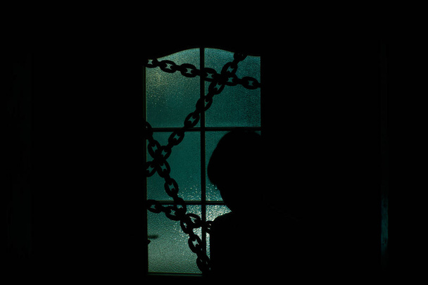 超自然的な緑色の光のチェーンとガラスドアの後ろの子供の暗いシルエット。ハロウィーンの鎖のドアの後ろの部屋に 一人で閉じ込められてた夜の誘拐家の中の悪お化け屋敷の中. - 写真・画像