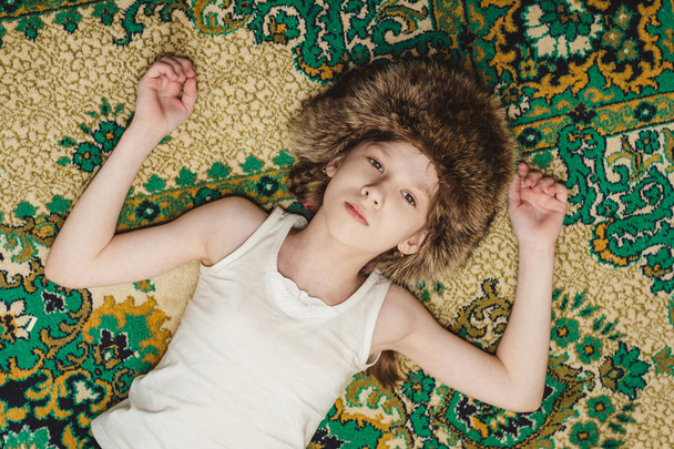 Ein kleines Mädchen mit Pelzmütze liegt auf einem alten Textilteppich. Kind entspannt mit geschlossenen Augen. - Foto, Bild