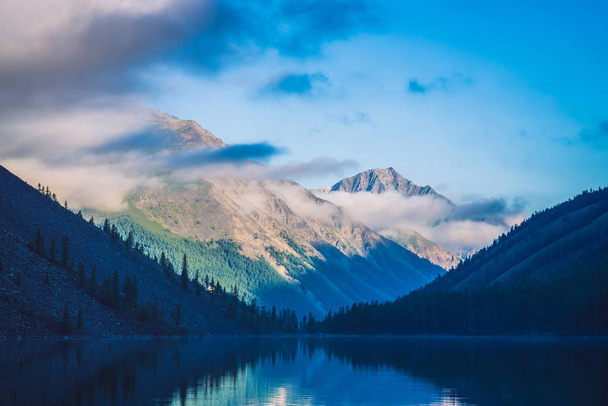 Amazing blue silhouettes of mountains under blue cloudy sky. Beautiful ripples on water of mountain lake. Low clouds before mountain ridge. Wonderful highland landscape. Picturesque mountainscape. - Foto, Imagem