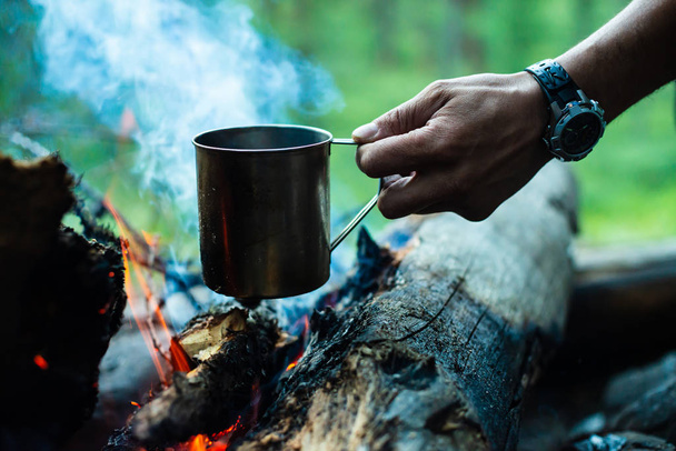 男はたき火の上に金属マグで茶を加熱しています。自然の上でホットドリンク。外風でお茶を飲む。人間の手にスチールマグカップ。夕暮れ時のキャンプ。夕暮れ時のロマンチックな暖かい雰囲気の屋外.アクティブな休息. - 写真・画像