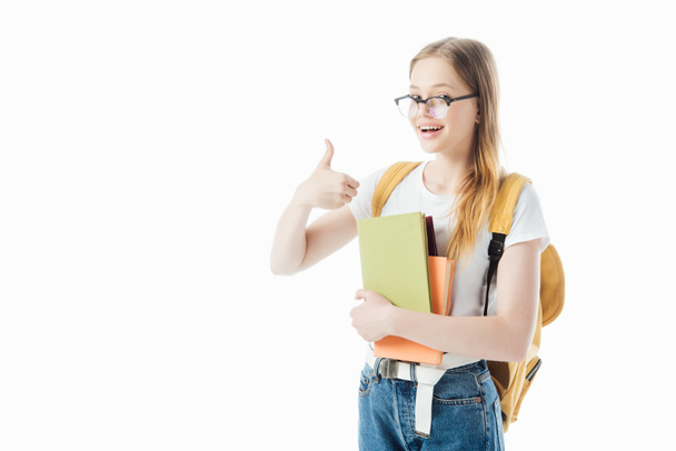 smiling schoolgirl with backpack holding books and showing thumb up isolated on white - Fotoğraf, Görsel