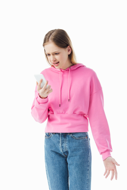 chica adolescente irritada en sudadera con capucha rosa y jeans sosteniendo teléfono inteligente aislado en blanco
 - Foto, Imagen