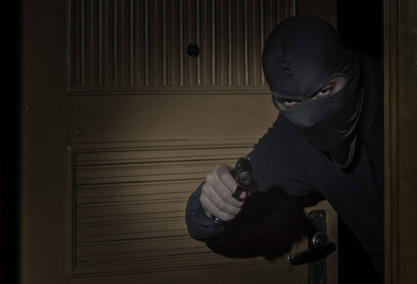 Ladrão com uma arma na mão e em um chapéu com uma fenda para os olhos foge para dentro da casa através da porta à noite ao luar
 - Foto, Imagem