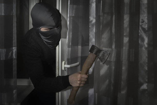 Thief with an ax in his hand and in a hat with a slit for the eyes sneaks into the house through the door at night in the moonlight - Photo, Image