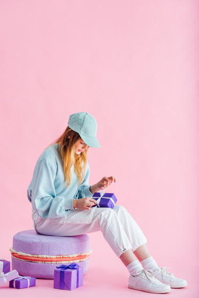pretty teenage girl in cap sitting on decorative macaroon and unpacking presents on pink - Photo, image