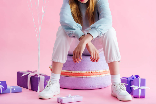 cropped view of teenage girl sitting on decorative macaroon near presents on pink - Фото, изображение