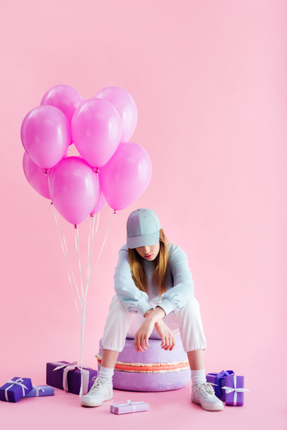 teenage girl in cap sitting on decorative macaroon near presents and balloons on pink - Foto, imagen