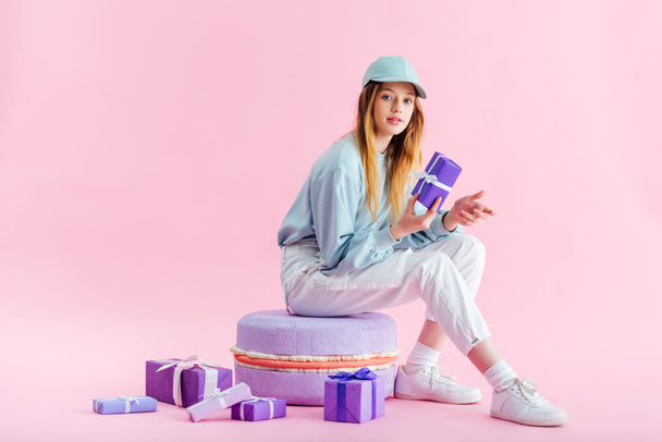 teenage girl in cap sitting on decorative macaroon near presents on pink - Photo, Image