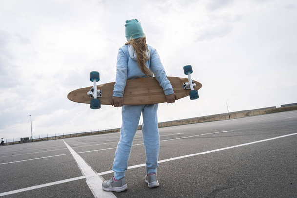 A girl in a hat and a blue sweater is holding a large longboard. - Zdjęcie, obraz