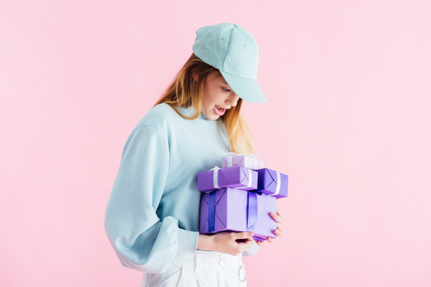 excited pretty teenage girl in cap looking at purple gifts isolated on pink - Φωτογραφία, εικόνα